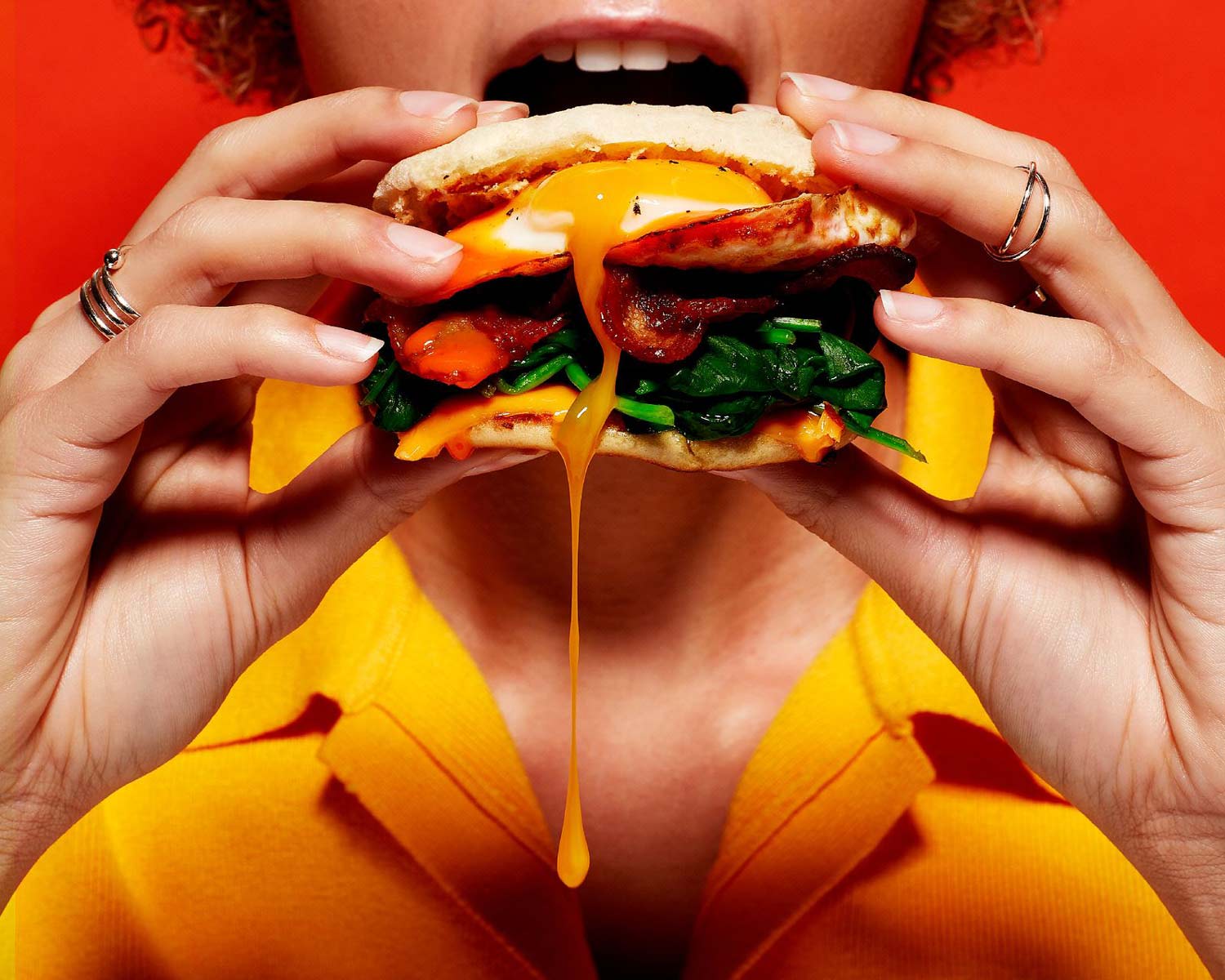Person holding a breakfast sandwich with spinach, bacon, melted cheese, and a runny egg yolk dripping down.