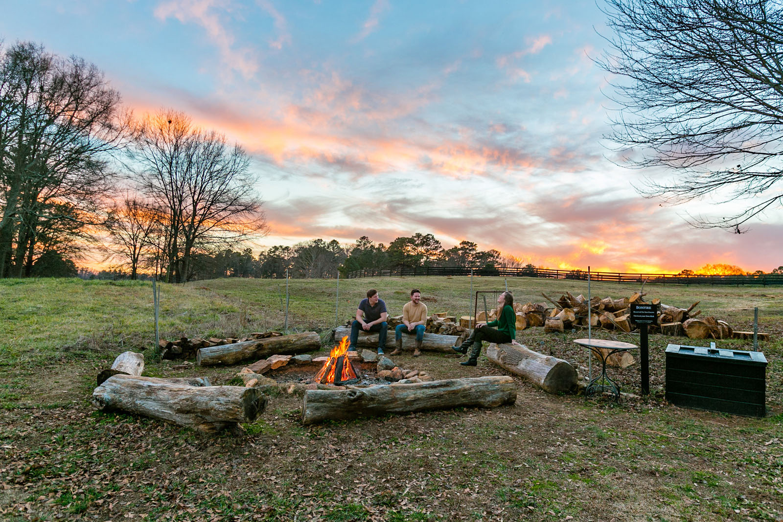 There are three smiling and laughing people sitting on long seats around a small bonfire at an open campsite at The Inn at Serenbe in Chattahoochee Hills, Georgia. There are trees scattered around them and in the background. The sky above is a light blue, scattered with the whispy orange-and-pink clouds of a recent sunset.