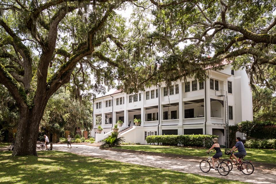 The white Greyfield Inn in Cumberland Island, Georgia, stands stately and stout next to a busy bike and walking path. The house is multi-storied with several windows on each level, with a staircase leading up to the front door. There are several people walking and biking on the path in front of the house. There are towering trees around the house with leaves so full the sky above is nearly blocked in the photo.