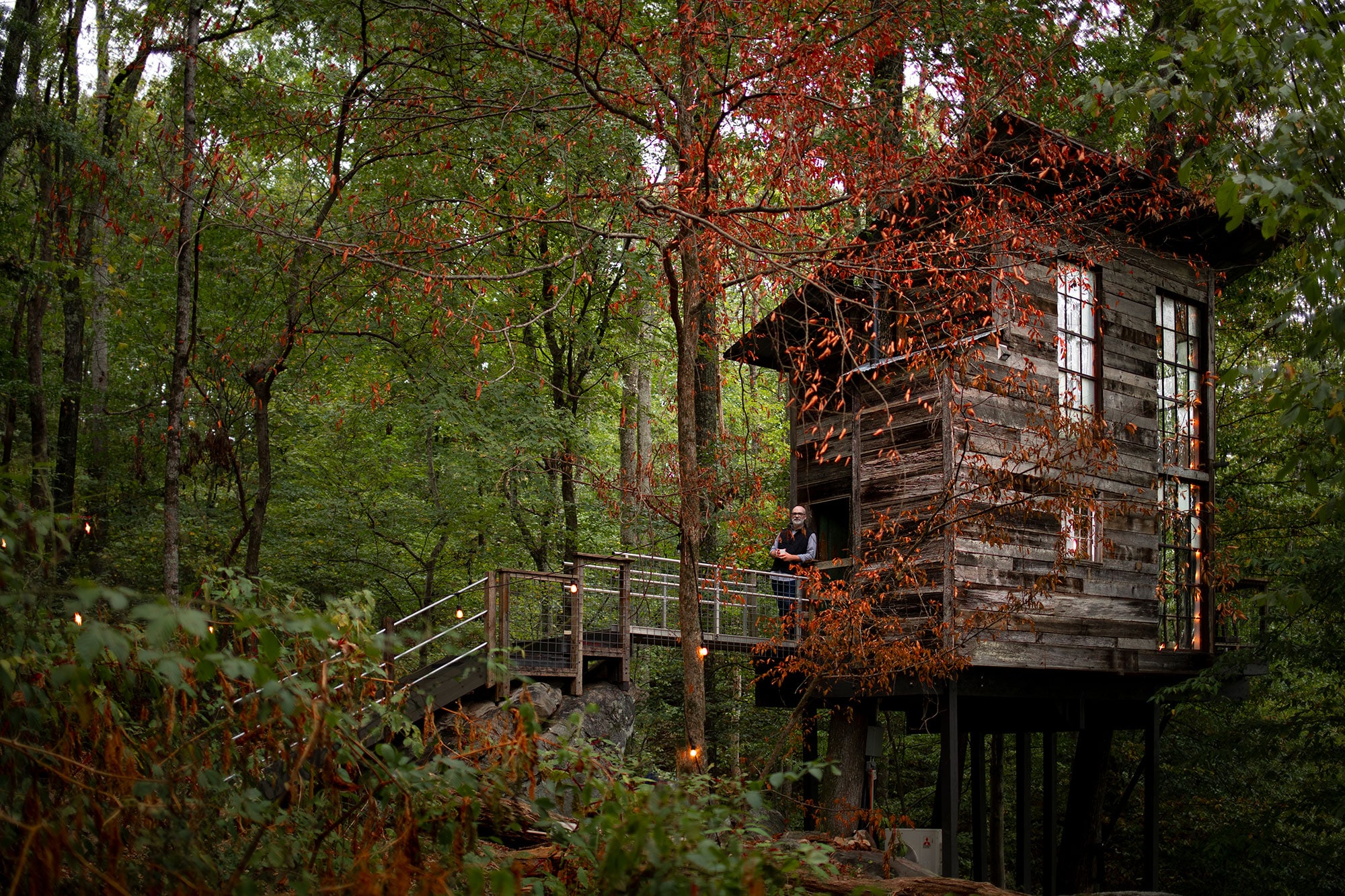 A modern, sleek-looking wooden treehouse stands on stilts amid deep, dense forestry at Treetop Hideaways in Flintstone, Georgia. A person stands on the bridgeway leading up to the door to the treehouse, leaning on the rails, smiling at the camera. There is a tall, red-leafed tree in front of the cabin, with its leaves just sparse enough to see through them clearly. There are twinkling dangling lights all around the scene.