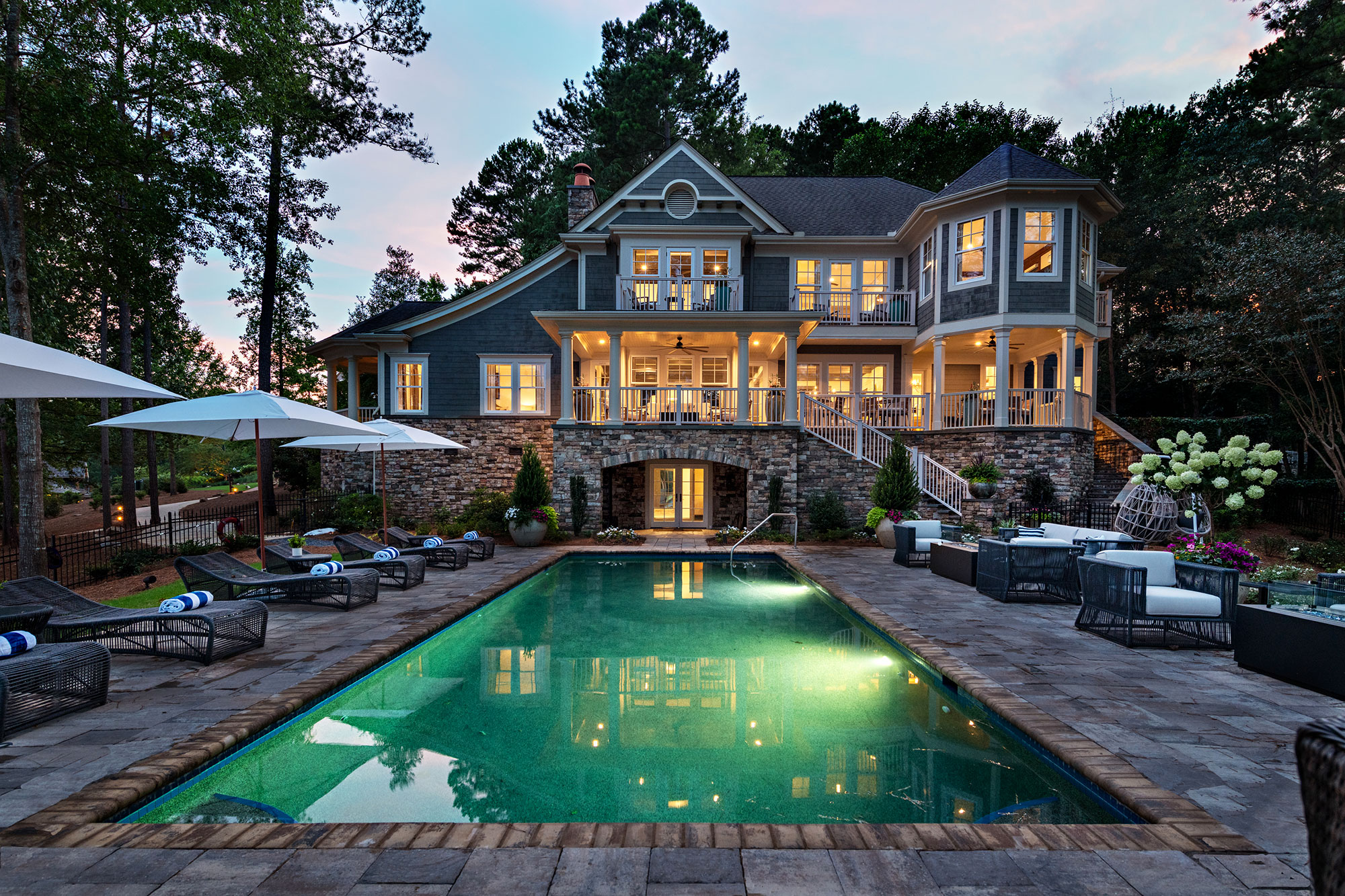 A magnanimous, multi-storied, stone-and-wood, mansion-looking house with a pool at the Ritz-Carlton Reynolds, Lake Oconee in Greensboro, Georgia, sits stately in the middle of the photo. It's dusk, with the sky above painted a dull blue-and-pink from the sunset. The windows are lit up in gold with the lights from the interior. There are tall pine trees all around the houe, and the pool is flanked by pool furniture and umbrellas.