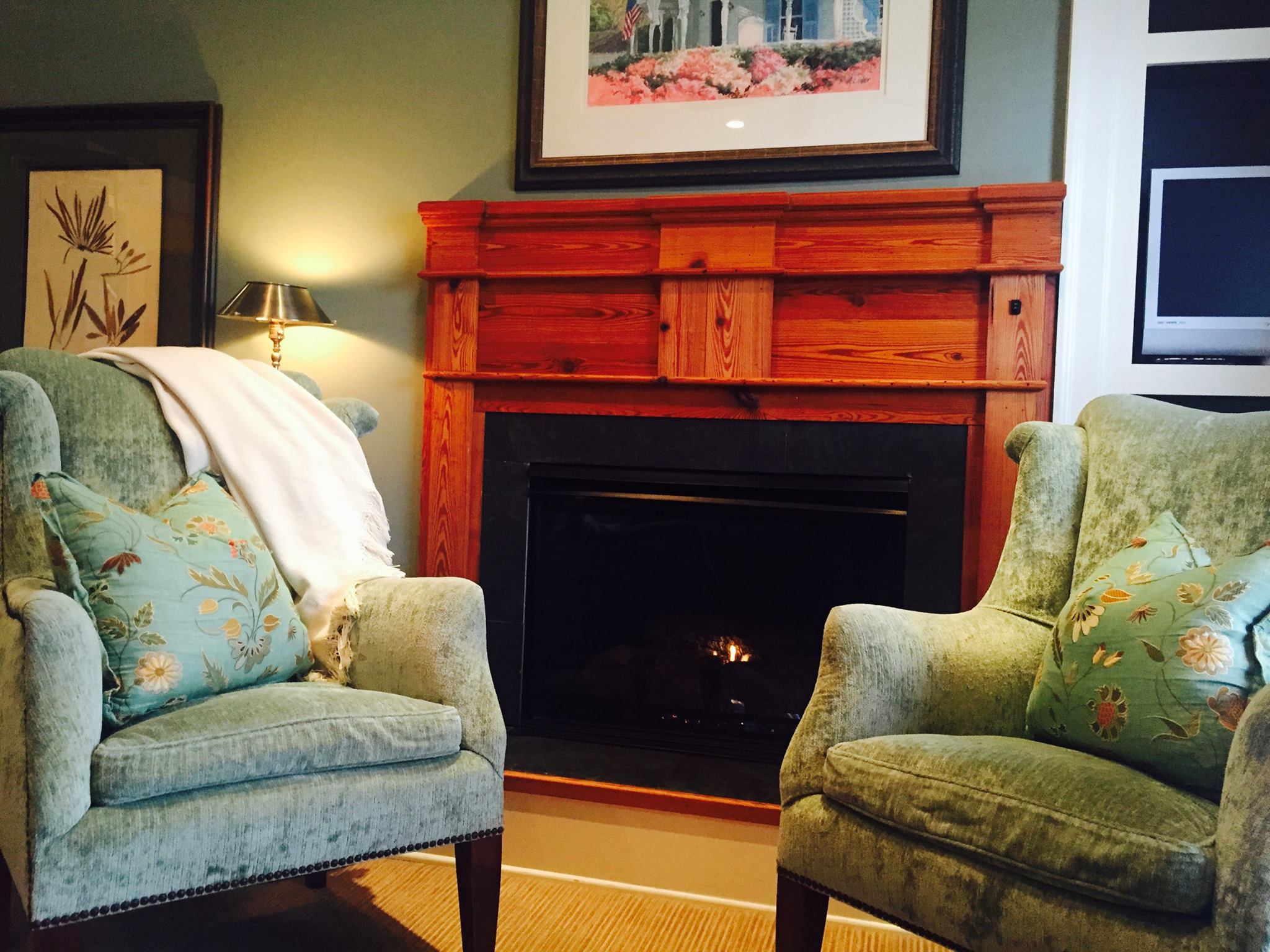 The interior of a cozy room at the James Madison Inn in Madison, Georgia. Two green, velvet armchairs sit next to each other, with a lit fireplace between them. There are framed pieces of art hung on the walls around the chairs.