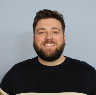 Smiling man with short brown hair, beard, nose ring, and black and beige shirt against a plain gray background.