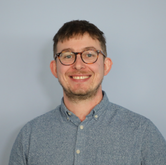 Smiling man with short brown hair, glasses, and a light blue button-up shirt standing against a plain light gray background.