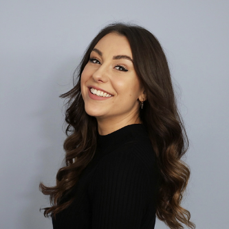 Smiling woman with long wavy brown hair wearing a black top against a light gray background.