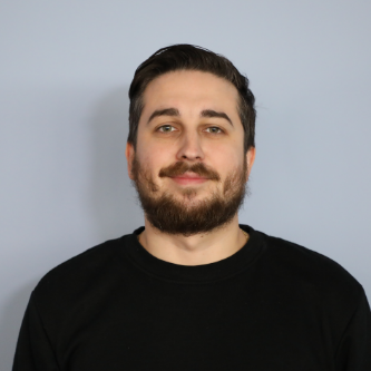 Portrait of a man with dark hair and beard wearing a black shirt against a gray background.