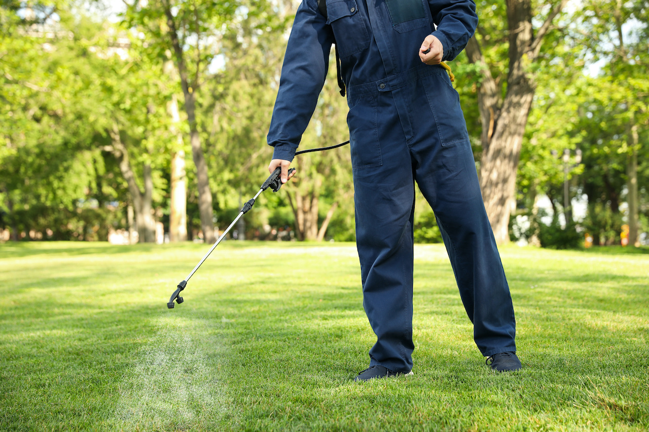Close-up of a lawn care worker’s gloved hand holding a spray wand, applying pesticide in a fine mist onto a green lawn outdoors.