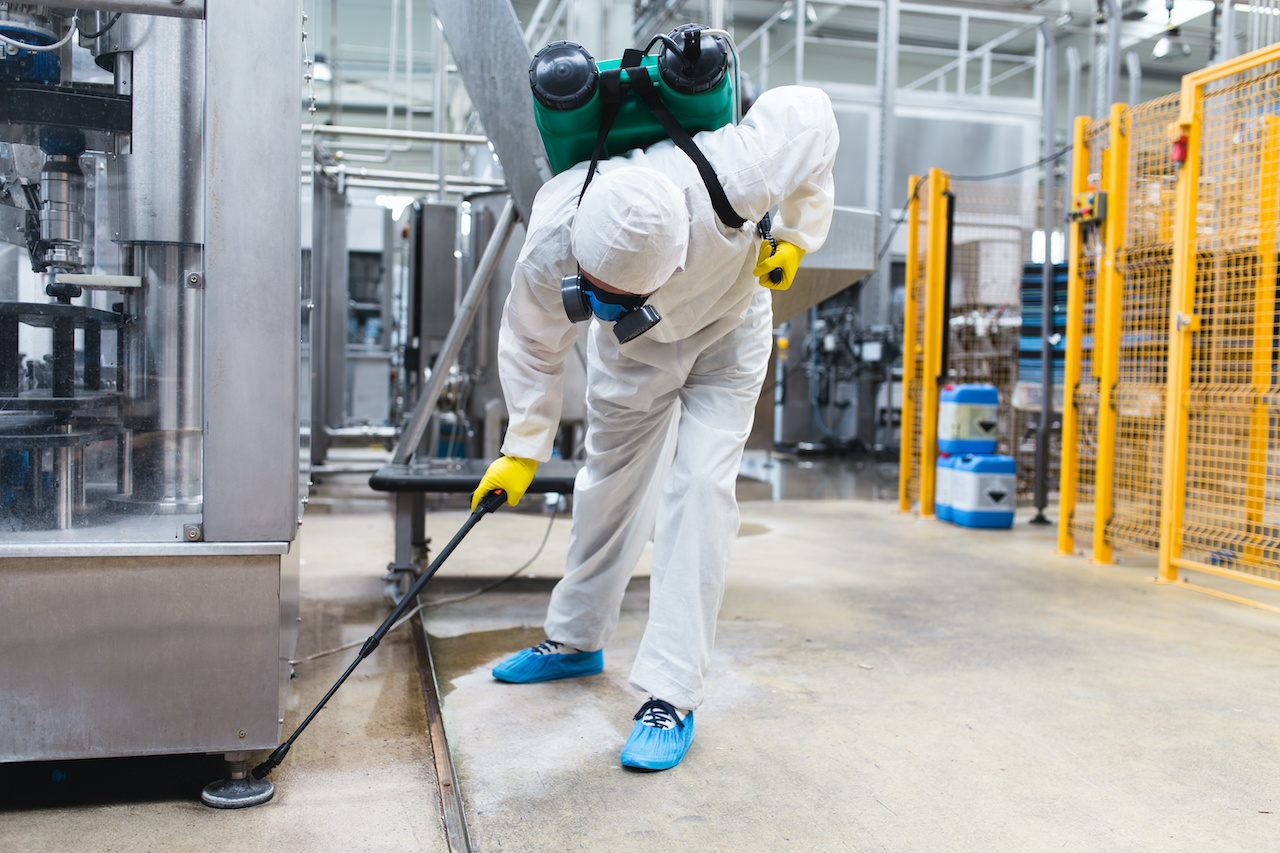 A pest control technician wearing a full-body protective suit and respirator holds a spray wand inside a factory or production facility, targeting pest-prone areas near industrial equipment.