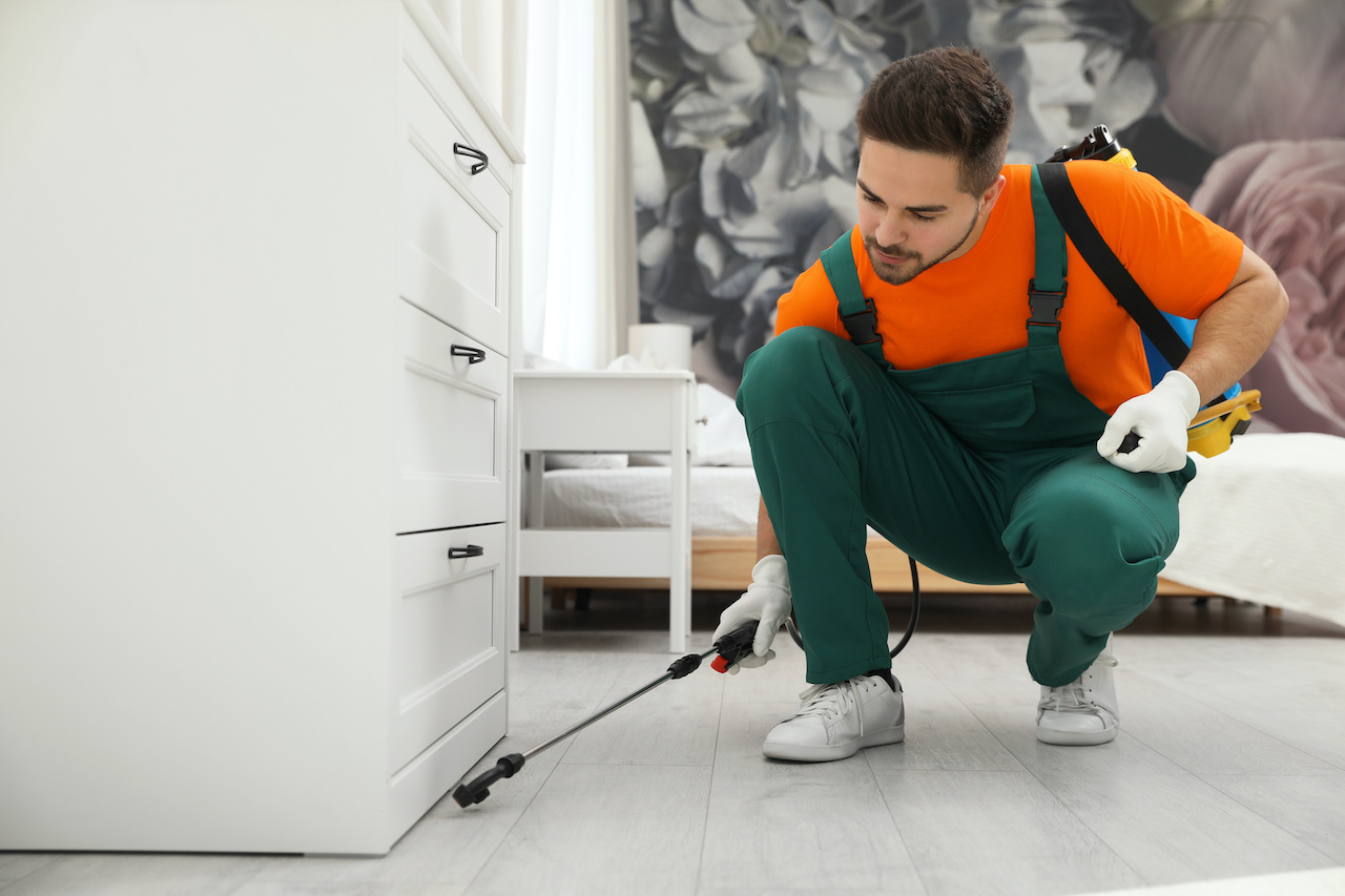 A pest control professional in an orange shirt and green coveralls crouches indoors beside a chest of drawers. He applies insecticide at floor level with a sprayer wand, targeting potential pest hotspots in a tidy room.