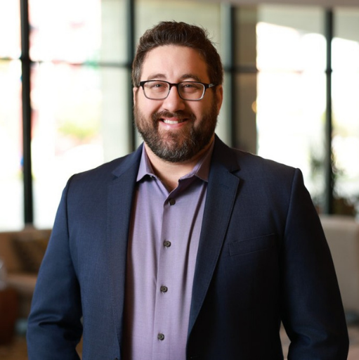 Smiling man with glasses and beard wearing a navy blazer over a checkered shirt against a blurred indoor background.