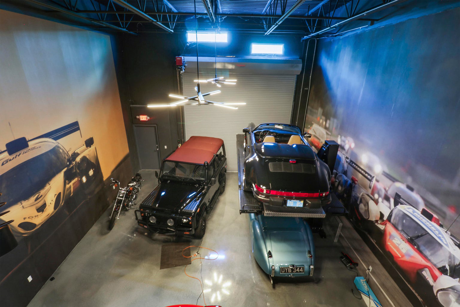 Indoor garage with a black SUV, a blue vintage car underneath a black convertible on a hydraulic lift, and a motorcycle, with large racing-themed wall murals.