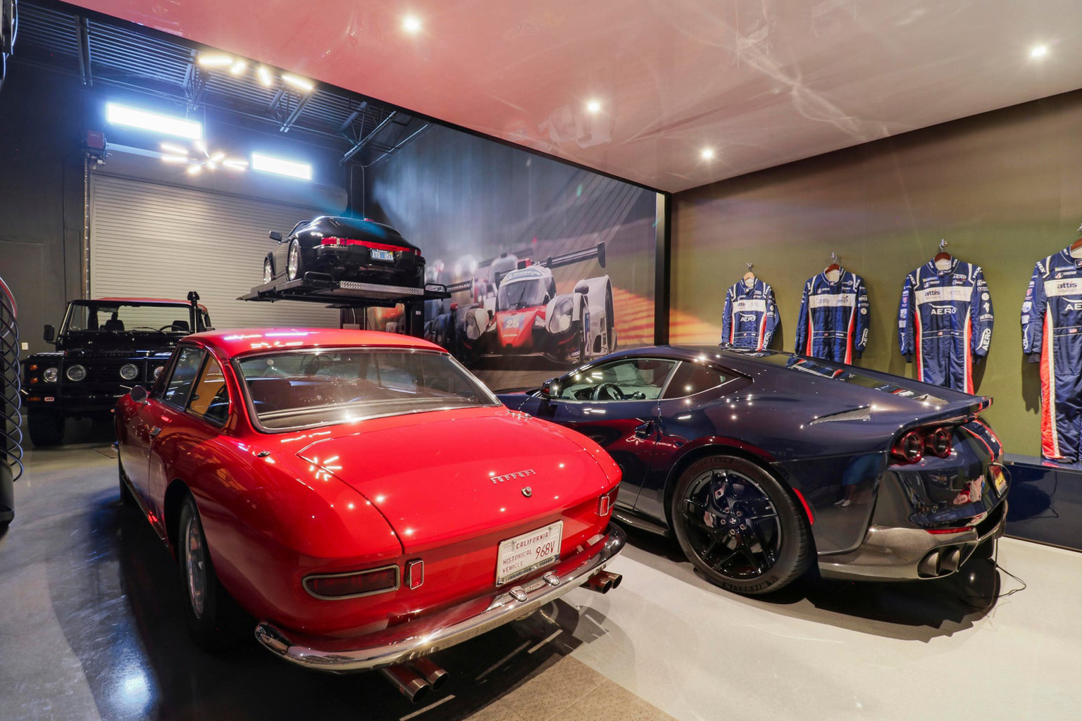 Interior garage with a red vintage Ferrari, a dark blue modern sports car, a black Land Rover, and a black Porsche elevated on a car lift, with racing suits displayed on the wall.