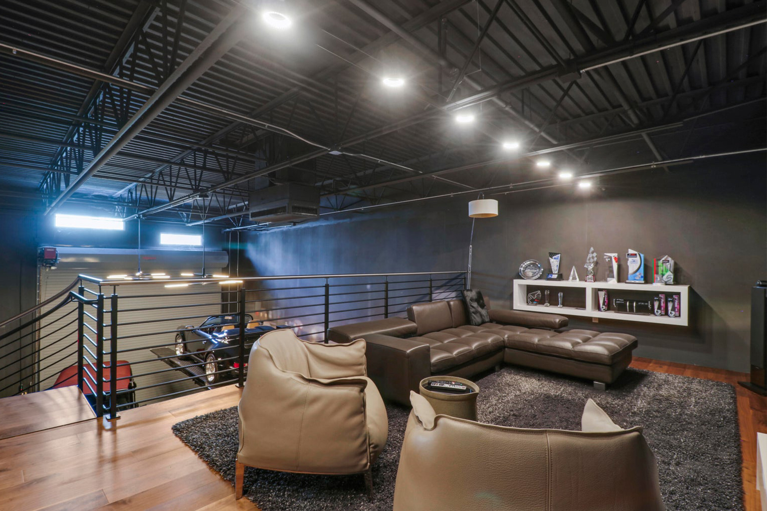 Modern loft living space with brown leather sectional sofa, two leather chairs, a gray rug, and a display shelf with trophies on a dark wall.