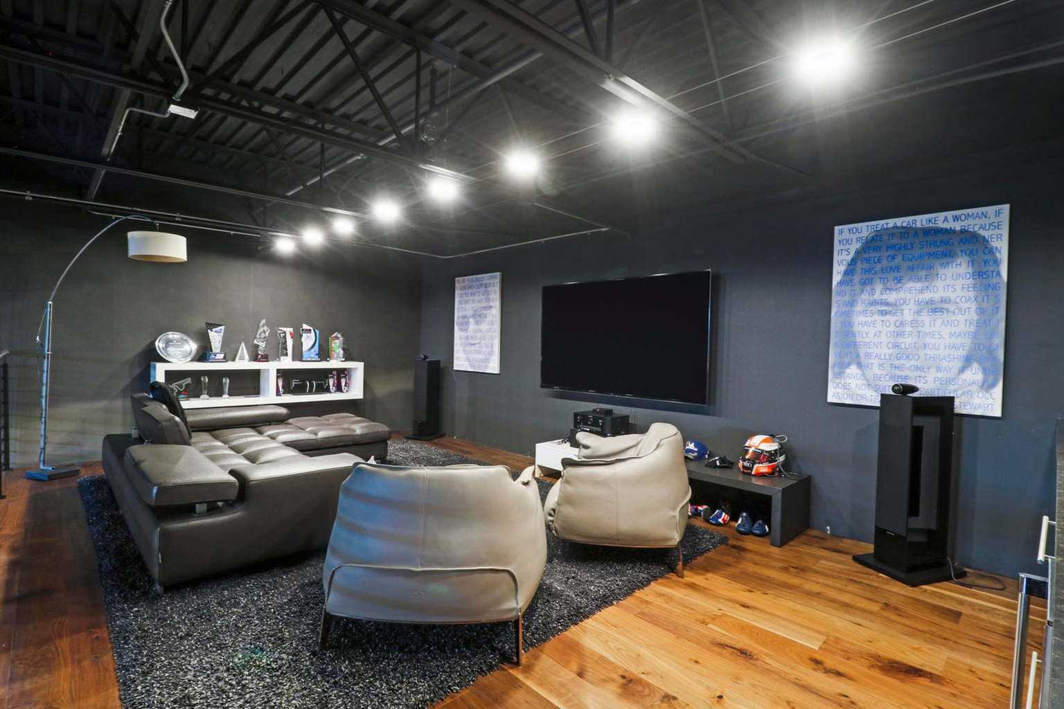 Modern living room with gray leather sectional sofa, two armchairs, large flat screen TV, and trophies displayed on a white shelf against dark walls with wooden floor.