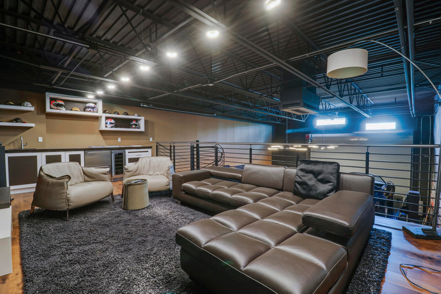 Modern lounge area with brown leather sectional sofa, two beige armchairs, a small round ottoman, and a kitchenette with display shelves holding helmets.