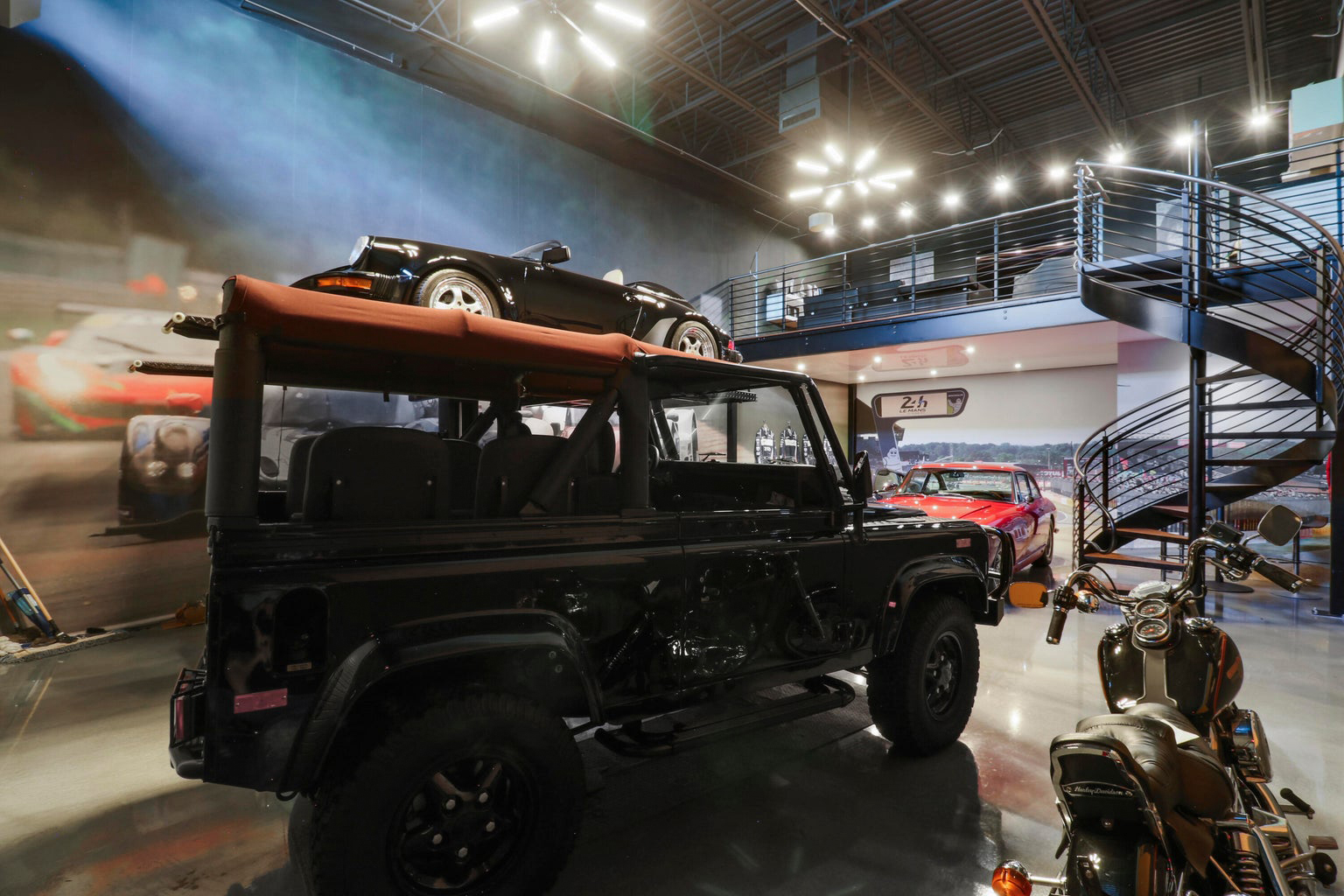 Interior of a modern garage with a black off-road vehicle carrying a small black convertible car on its roof, a motorcycle in the foreground, and a red classic car near a spiral staircase.