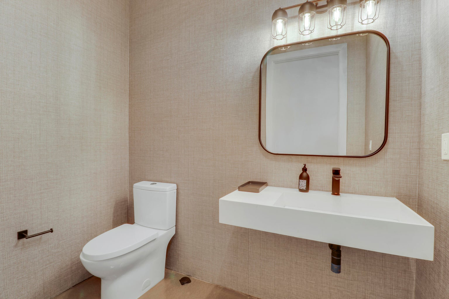 Modern bathroom with a white toilet, a floating white sink with brown faucet and soap dispenser, and a square mirror with rounded edges illuminated by four lights above.