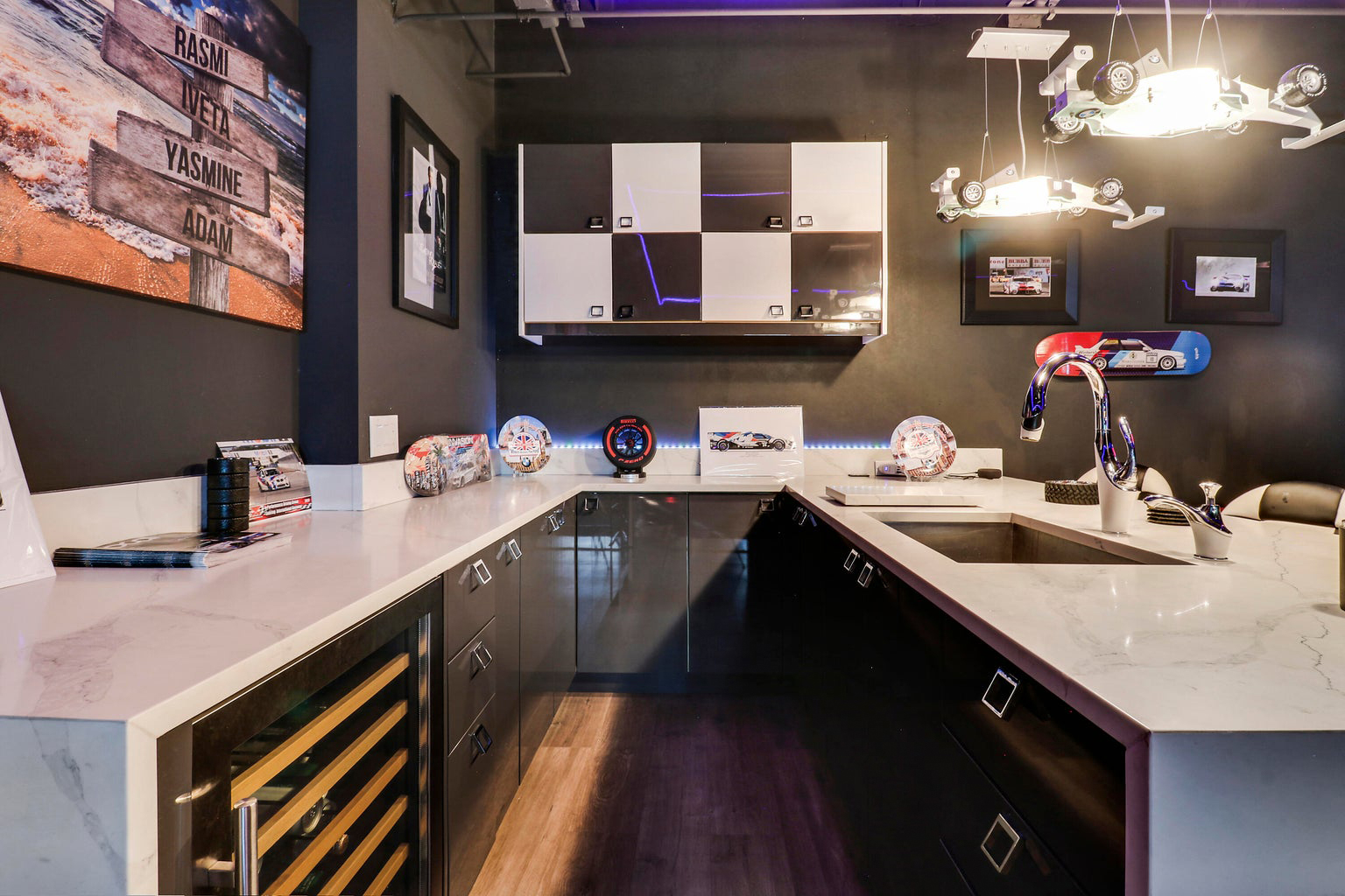 Modern kitchen with black and white cabinets, marble countertops, a stainless steel sink, racing car-themed decor, and hanging formula one car-shaped lights.