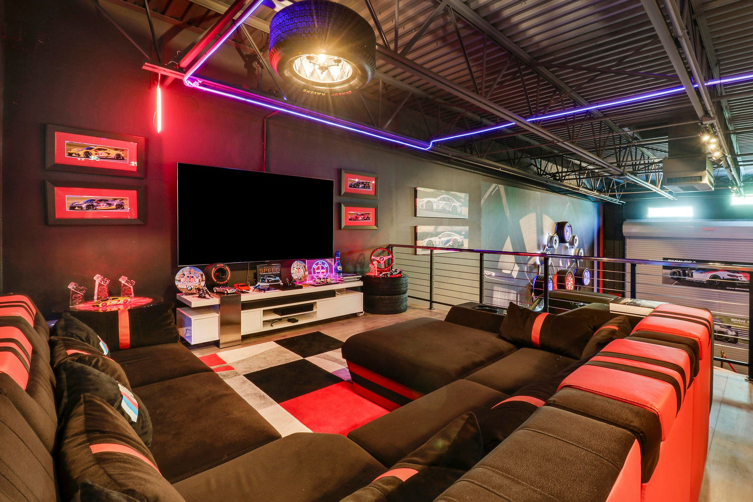 Modern lounge with large sectional sofa in black and red, mounted flat-screen TV, car-themed decor and neon lighting.