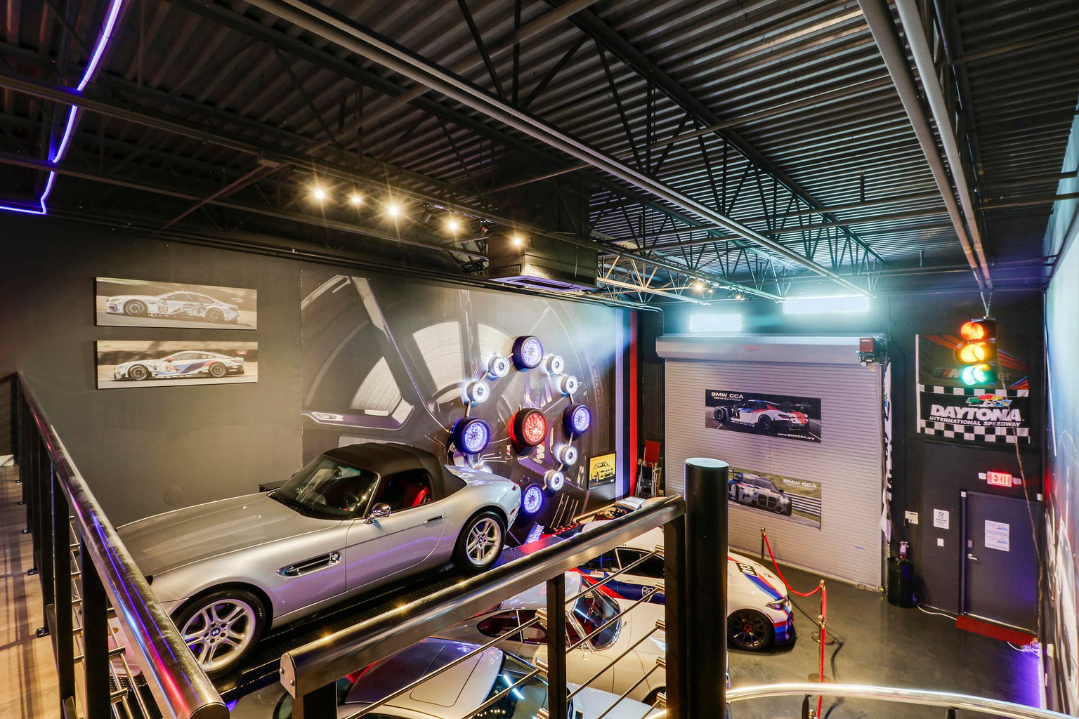 Indoor showroom with classic and race cars, featuring a silver convertible on a raised platform, illuminated wheel display on the wall.