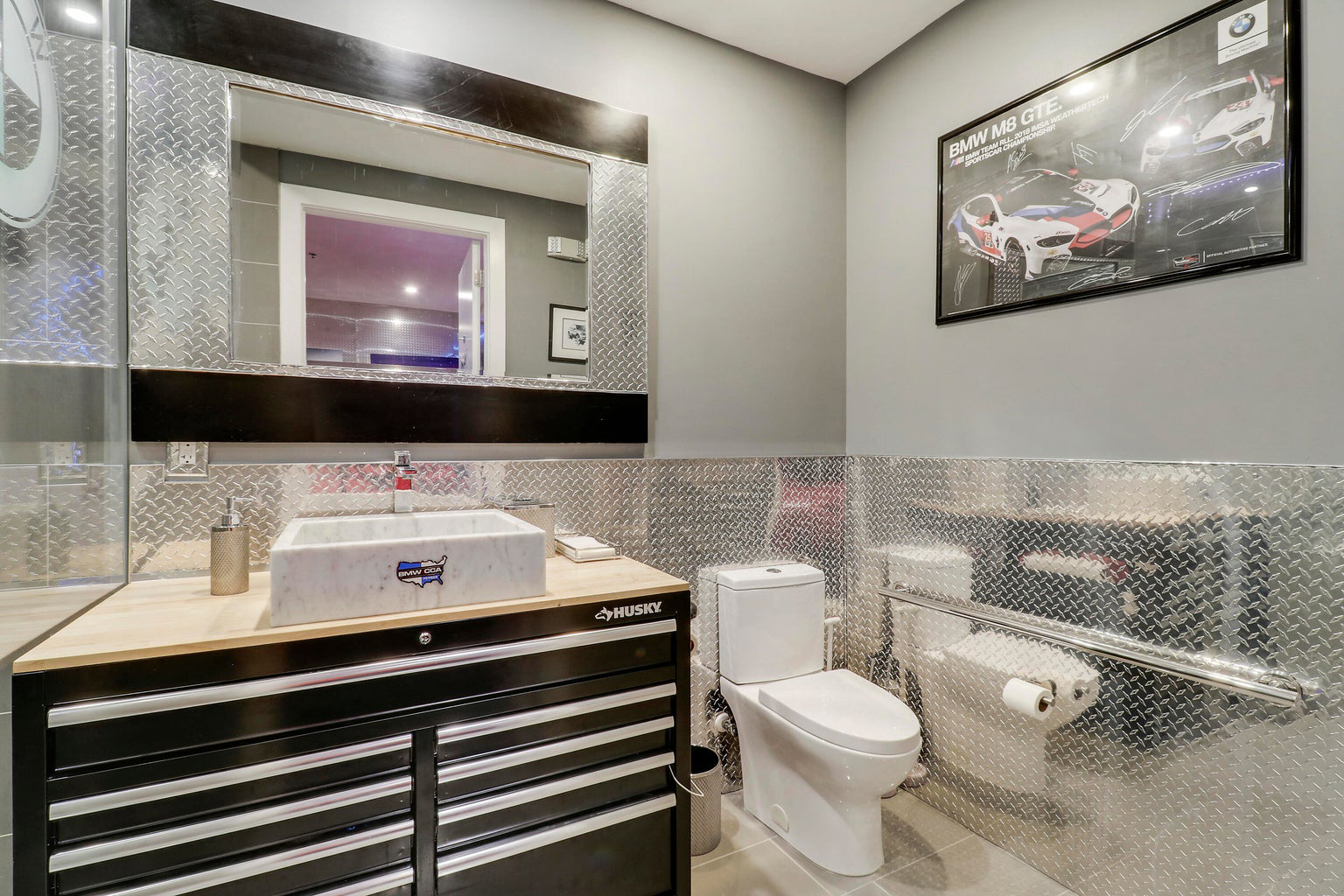 Modern bathroom with a white toilet, metal diamond plate wall paneling, a black tool chest vanity with rectangular marble sink, and a framed BMW M8 GTE poster.
