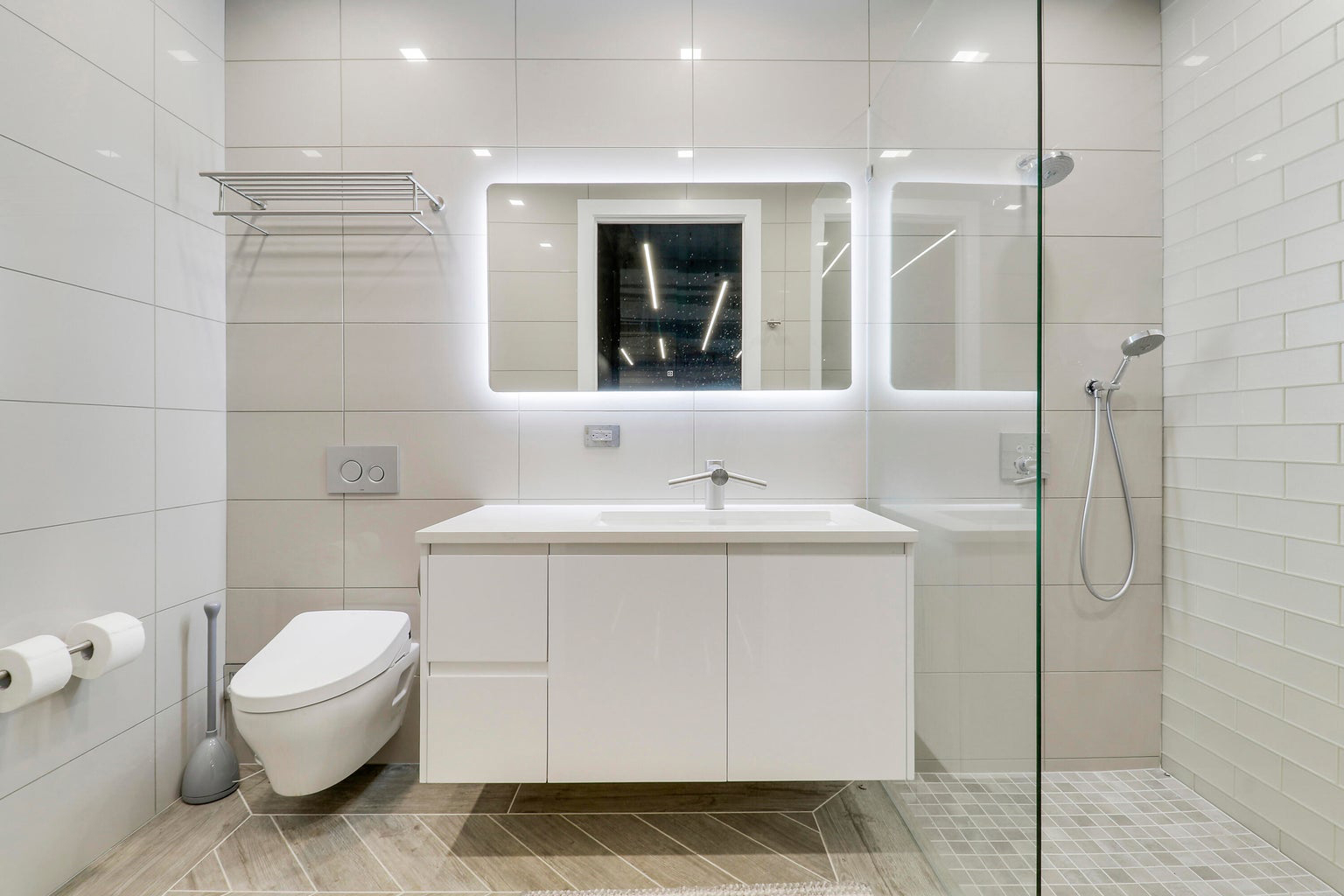 Minimalist modern bathroom with a wall-mounted toilet, floating vanity with sink, illuminated rectangular mirror, and glass-enclosed shower.