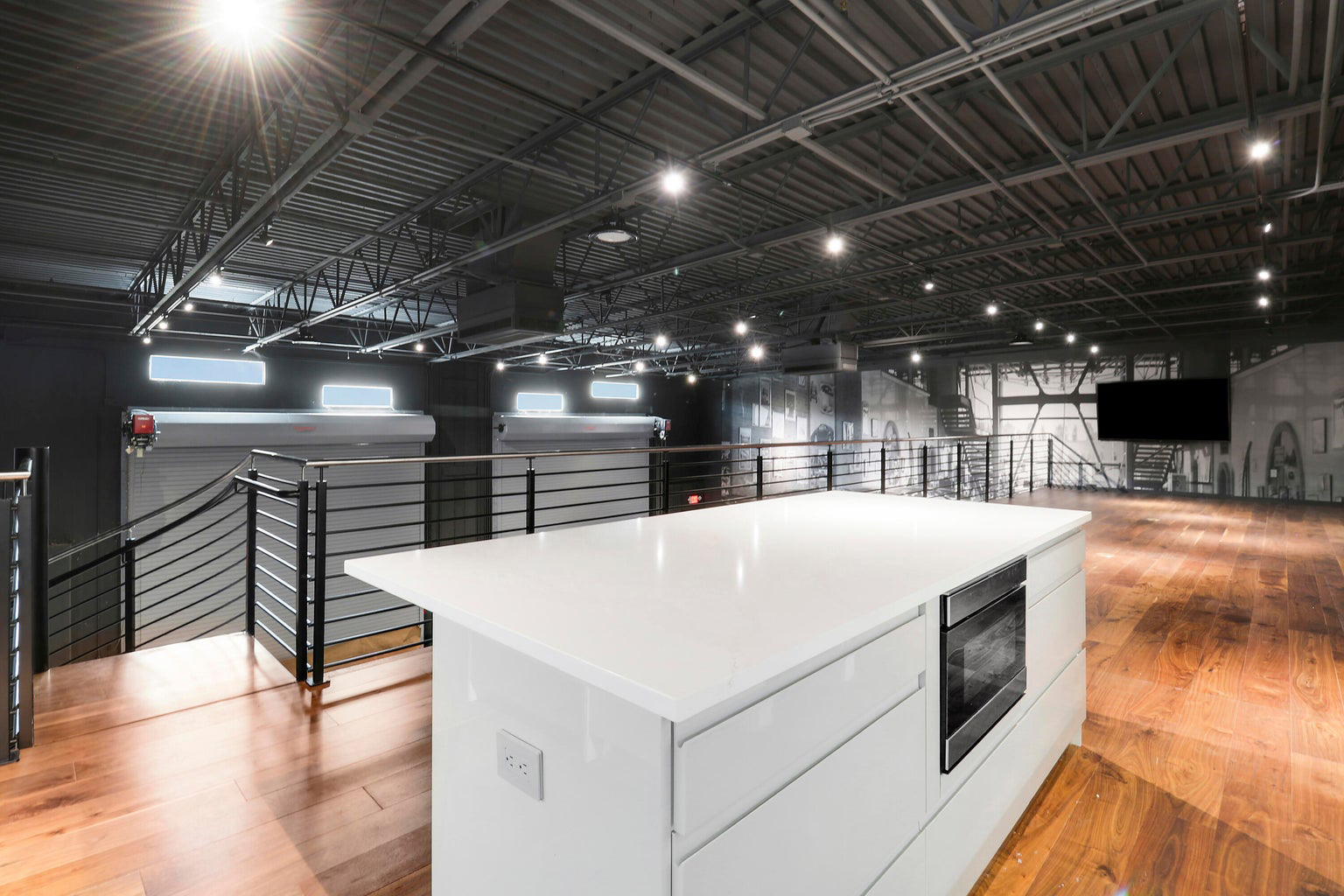 Modern industrial loft kitchen with white island countertop, built-in oven, wooden flooring, and metal railings overlooking large garage doors.