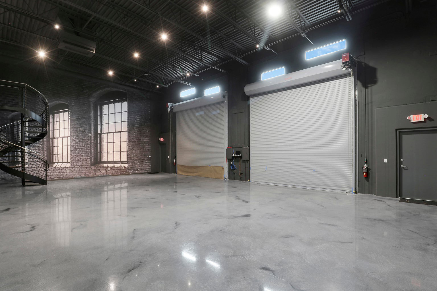 Spacious industrial warehouse interior with polished concrete floor, two large closed gray roll-up doors, and a black spiral staircase.