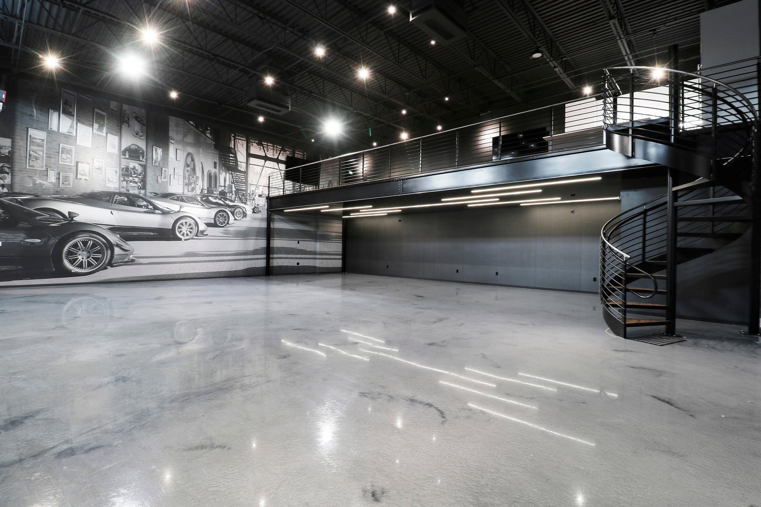 Empty modern garage space with polished concrete floor, black metal spiral staircase, mezzanine, and wall mural of luxury cars.