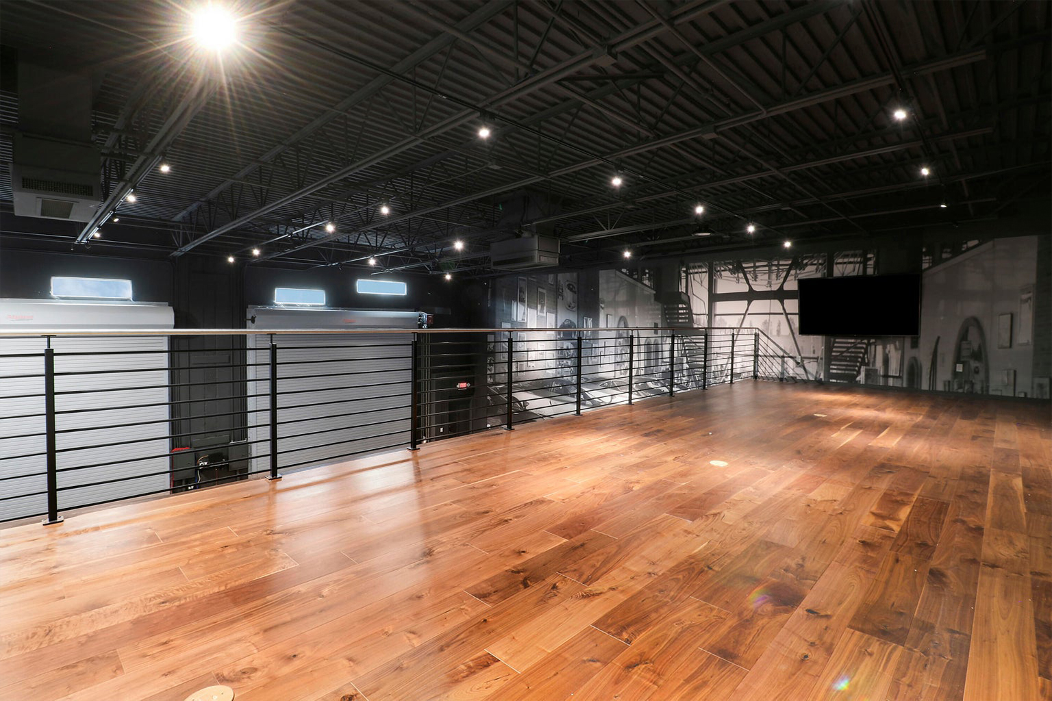 Spacious loft area with polished wooden floor, black metal railing, and industrial ceiling with exposed beams and lighting.