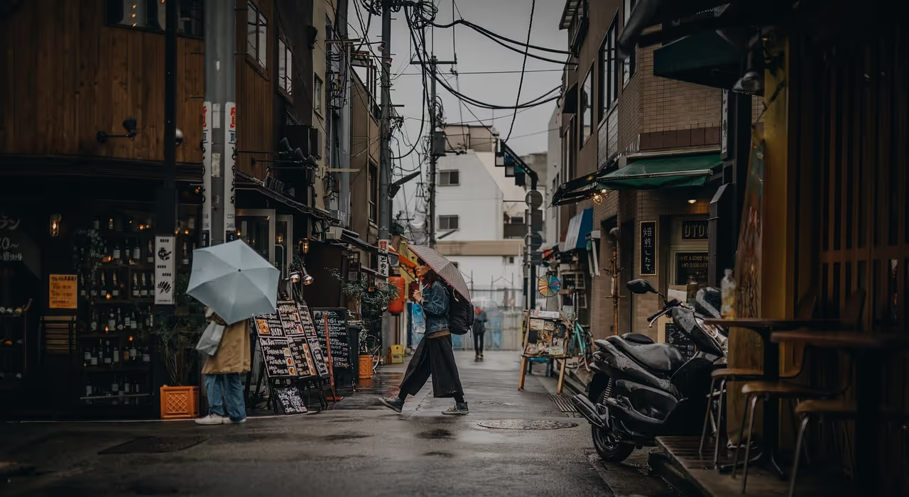 비 오는 날 젖은 포장도로가 있는 좁은 도시 골목. 우산을 든 두 사람이 작은 가게와 길가의 오토바이 옆을 지나갑니다. 머리 위로 전선이 교차하고 가게 창에서 은은한 빛이 나옵니다.