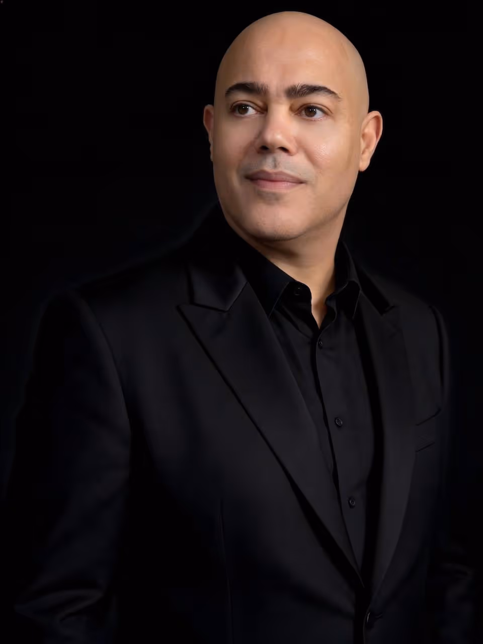 Portrait of a bald man in a black shirt and suit jacket looking slightly right on black background.