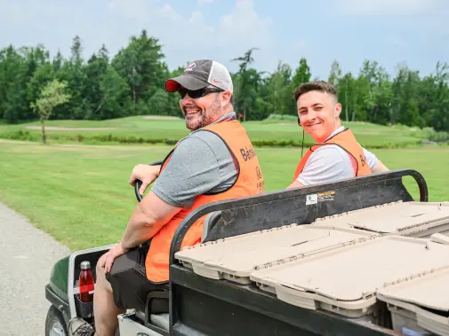 Deux bénévoles en gilet orange assis à l’arrière d’une voiturette de golf.