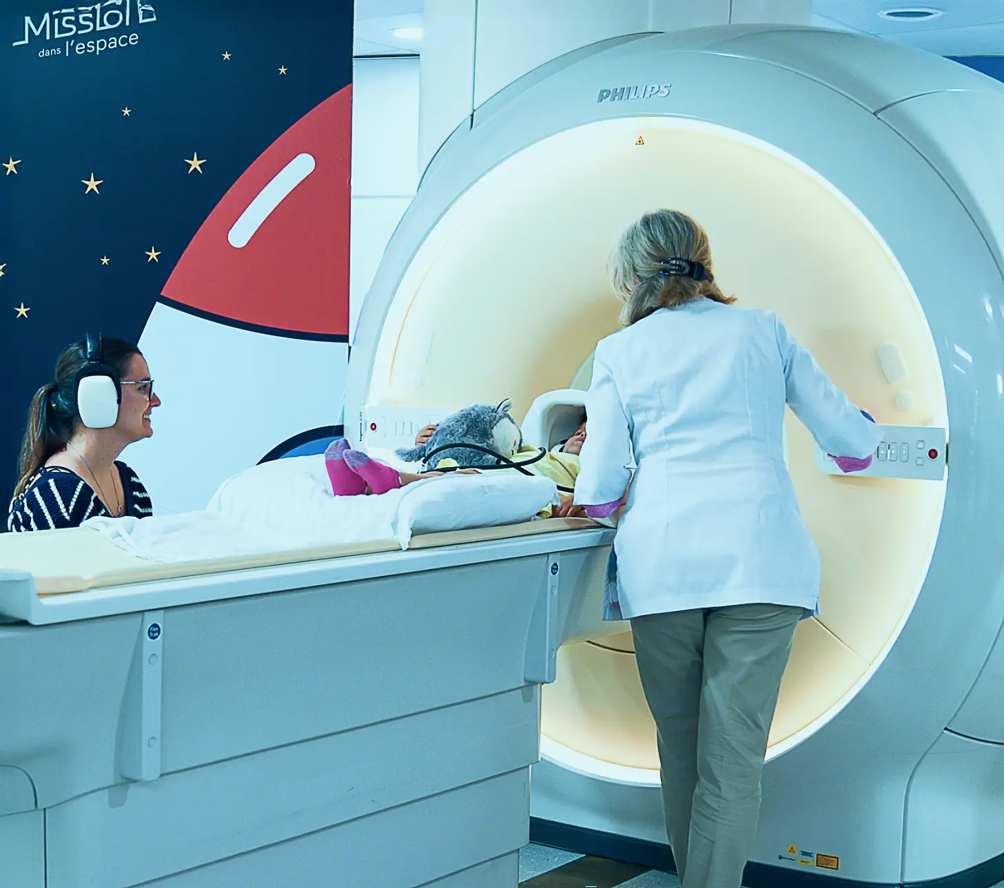 Enfant préparé pour un examen d’IRM pédiatrique au CHUS avec accompagnatrice et technologue.
