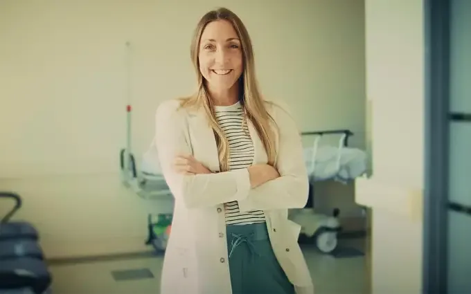 Médecin souriante en blouse blanche posant bras croisés dans un couloir d’hôpital.