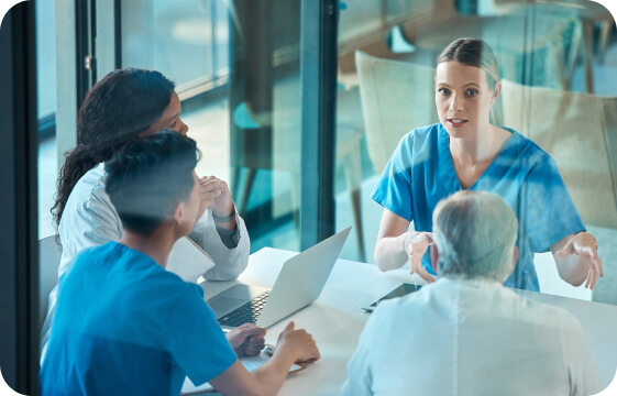 Medical professionals in blue scrubs and white coats discussing over a laptop in a meeting room.