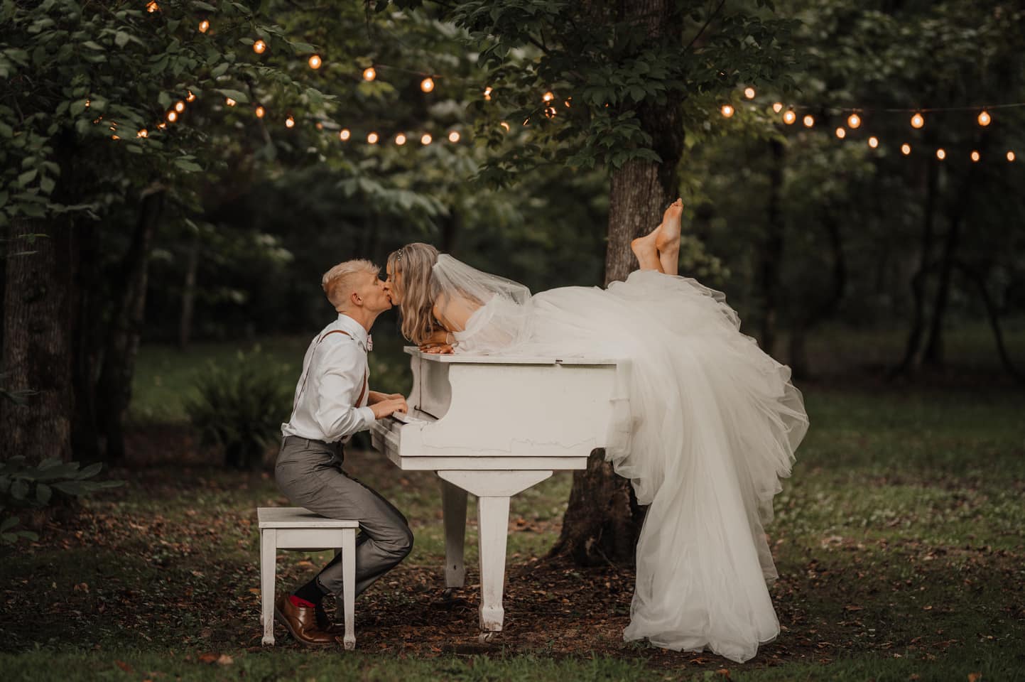Romantic wedding photo of bride laying across a white baby grand piano in the woods while her groom leans in for a kiss.  
