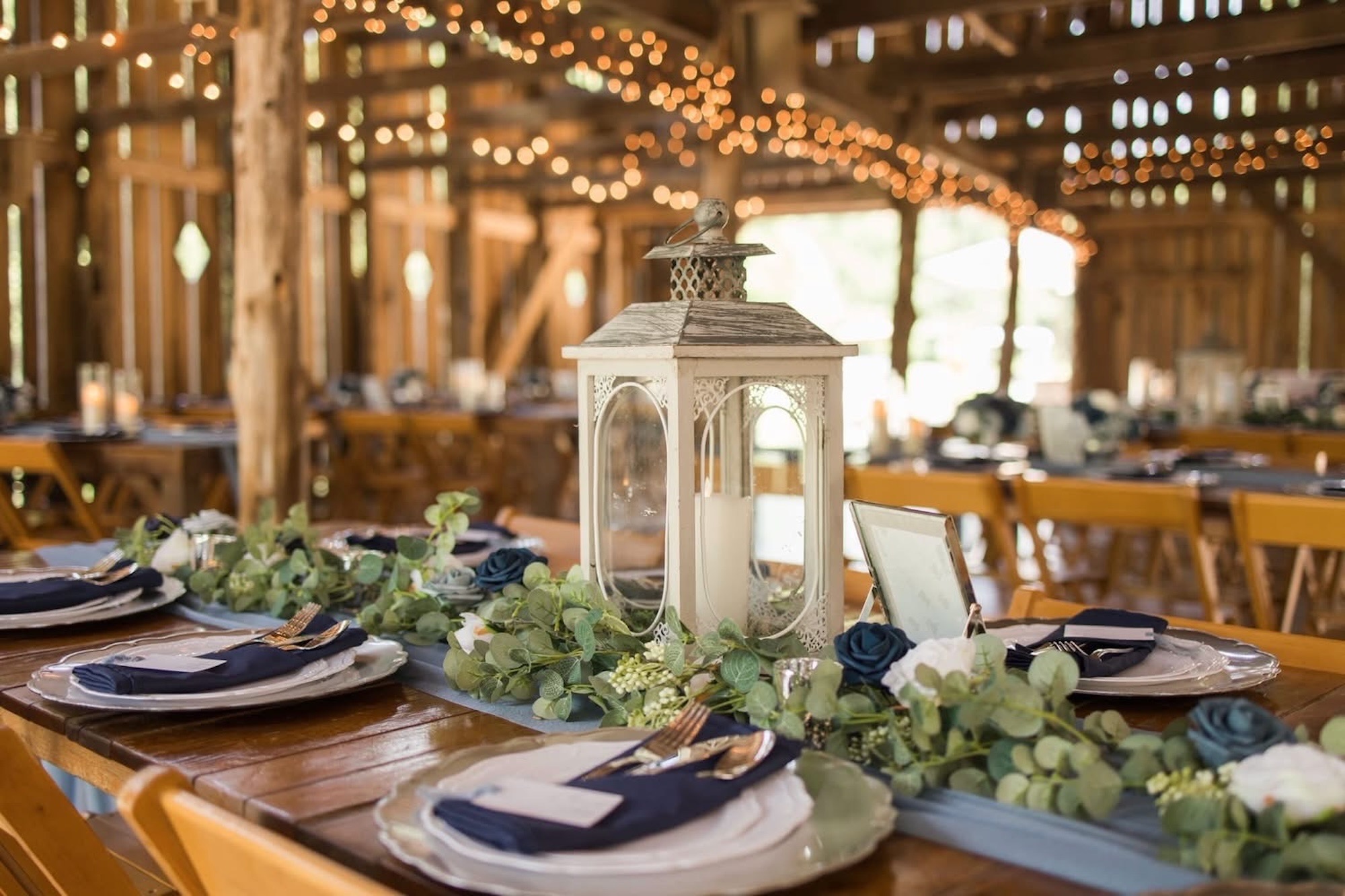 Wedding reception tablescapes feature wooden farmhouse tables decorated with white lanterns and greenery centerpiecesa and blue napkins