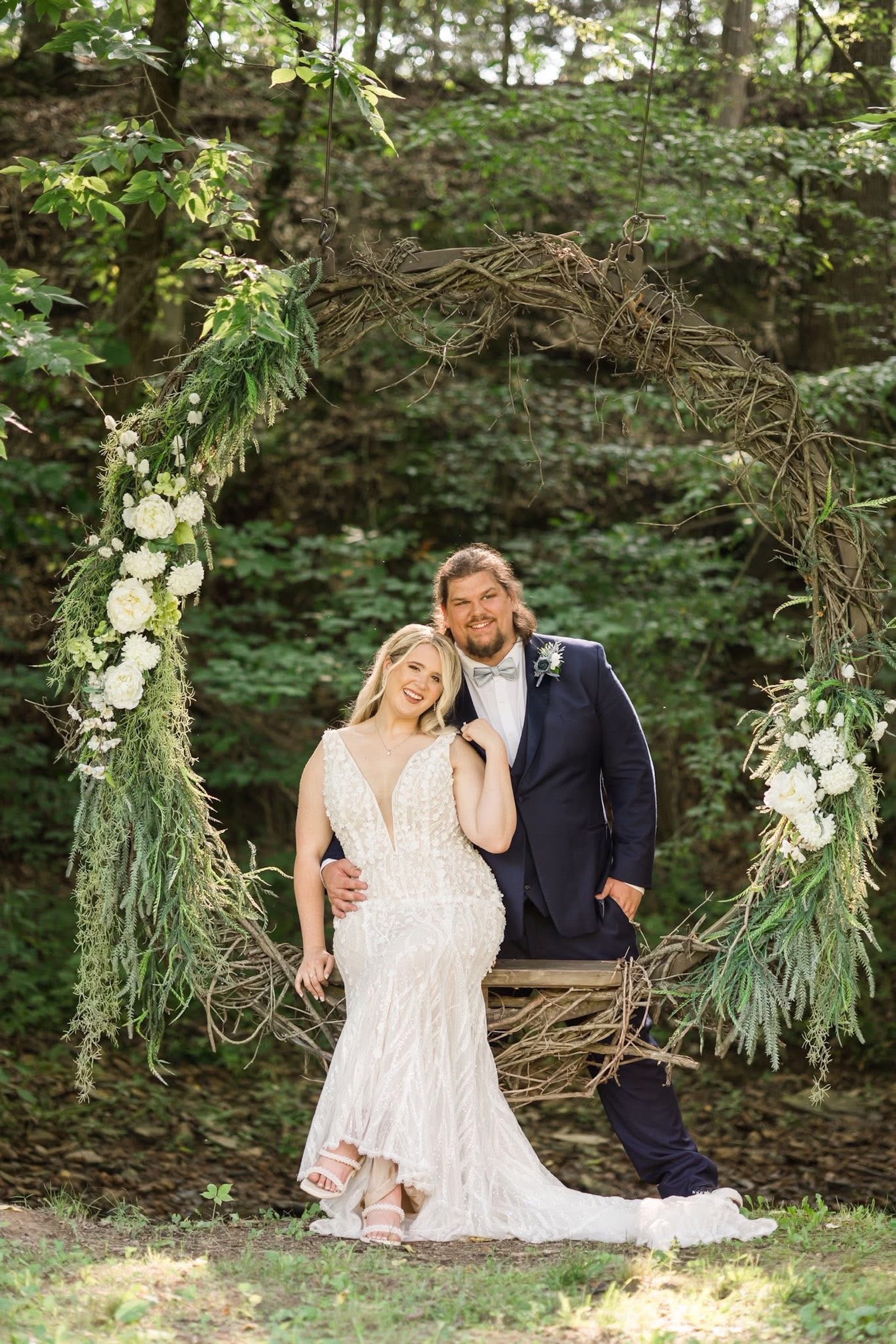 Bride sits on the circle grapevine swing adorned with flowers while the groom stands behind her.  One of the beautiful photo ops at The Barn at Cedar Grove 
