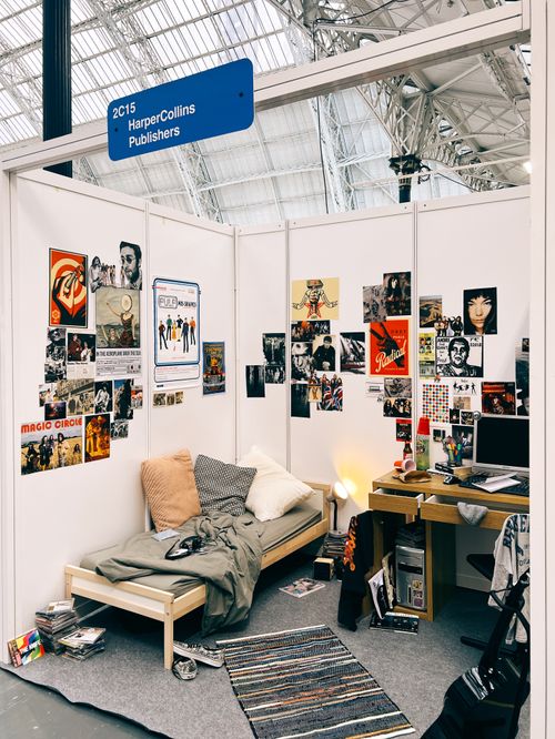 Trade show booth structure that has been turned into a dorm-style bedroom space, with bed and computer desk, soft furnishings and props such as posters on the walls, drinks cups and bottles and clothing strewn around the space