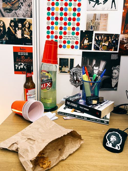 Section of a computer desk with wall posters visible in the background, drinks cups, bottles and Margarita cocktail mix, a croissant bag with crumbs, books, a pen pot with pens in it and a computer mouse
