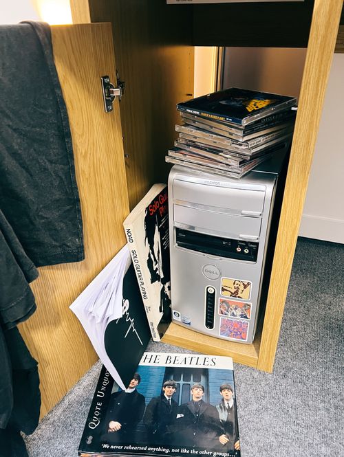 An opened cupboard in the lower section of a computer desk, with a computer modem tower visible. Mixed CDs and music books are scattered around it