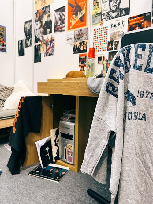 A perspective of a dorm-style room with posters and personal items strewn about is blurred in the background, and a Berkeley University t-shirt is hung over a chair in focus in the foreground