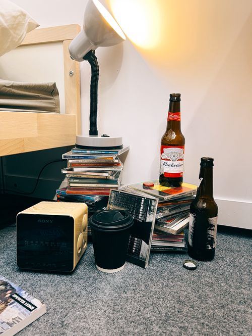Piles of mixed CDs sit on the floor next to a bed, with a desk lamp and empty beer bottles atop. An old fashioned alarm clock and coffee cup sit in front.