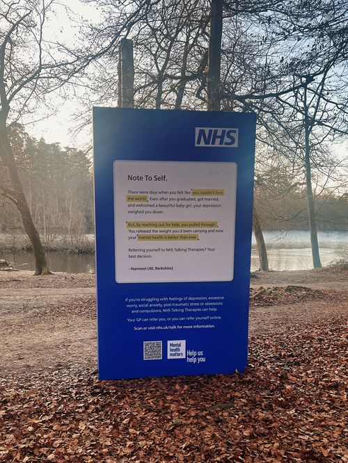 A freestanding blue box structure with a personal note and the NHS logo printed on the front of it stands in a park-like environment, leaves scattered around it on the ground. Trees and a lake can be seen in the background.