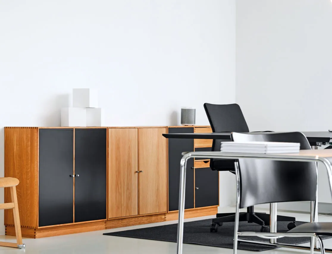 Modern office interior with wooden cabinets featuring black and natural wood doors, a black office chair, a table, and a stack of white books.