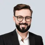Smiling man with glasses and a beard wearing a black suit and white shirt against a light background.