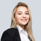 Smiling blonde woman in professional attire with white blouse and black blazer against a light gray background.