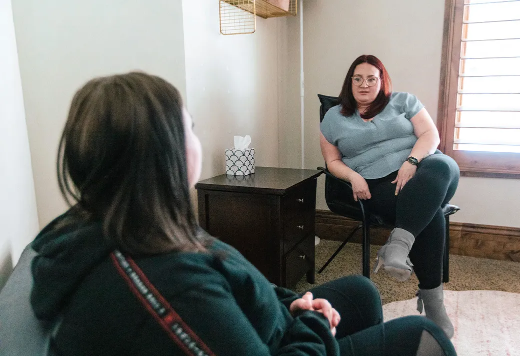 Two women sit and talk in a cozy room, one listening while the other speaks near a window.
