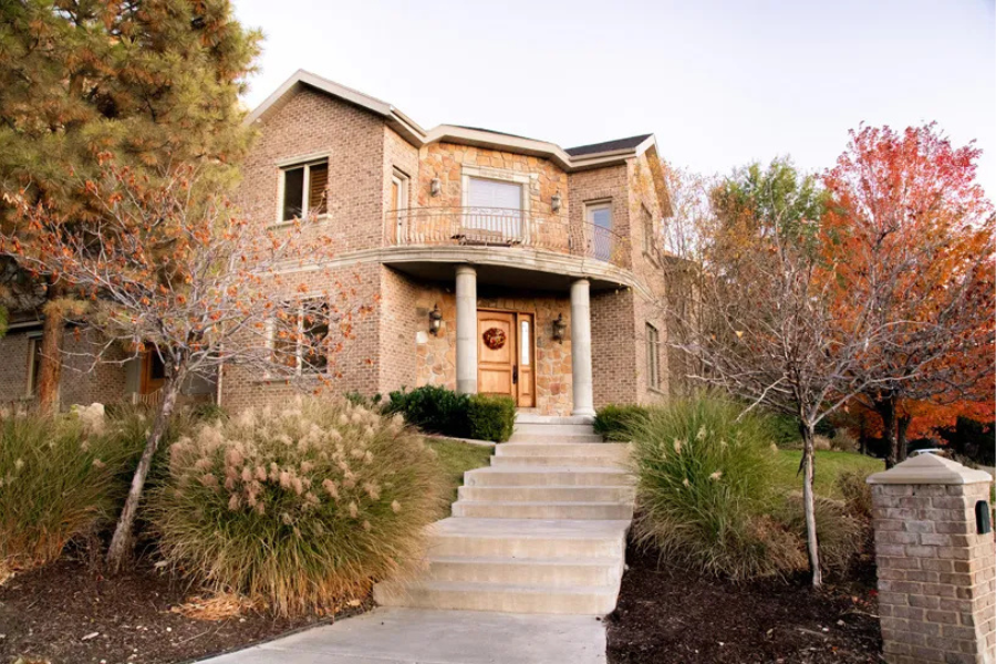 Two-story brick and stone residential building with a wooden front door, columned porch, and surrounding autumn foliage, viewed from the base of a front walkway lined with ornamental grasses and trees with colorful leaves.
